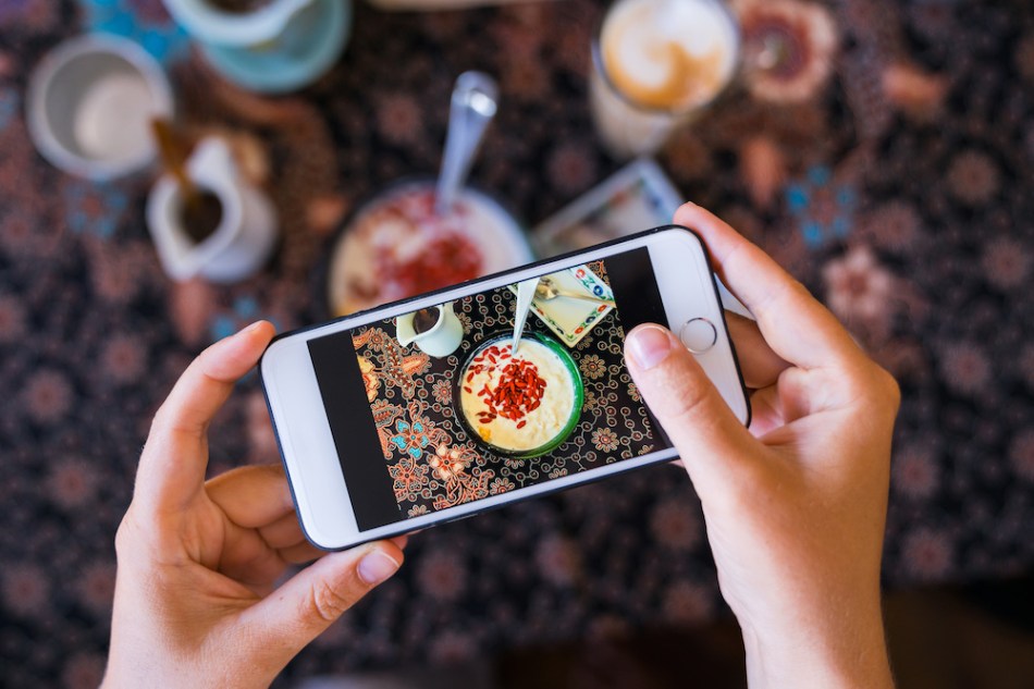 Hands with the phone close-up pictures of food. Breakfast, homemade oatmeal with fruit and goji berries.